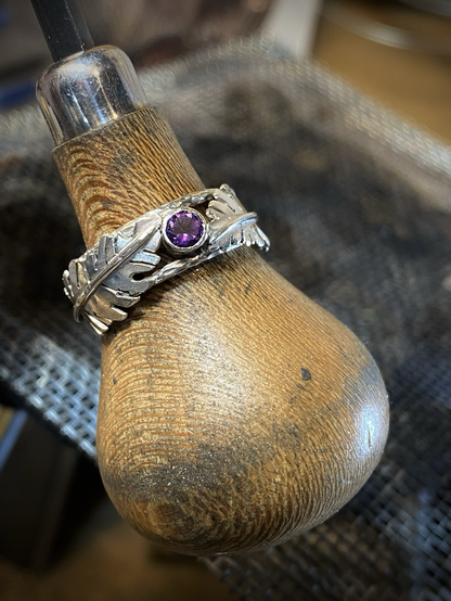 A close up view of the sterling silver ring from the previous photo being displayed on a stone setting tool. The handle of the tool being wooden shows waves of wood grain on its light colored wood surface. The tools handle has signs of use and wear, showing very small particles of silver dust and dirt from years of use. 