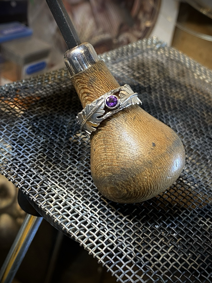 A recently finished sterling silver ring featuring a round juicy purple amethyst stone in the middle with two long leaves on either side in alternating directions. The ring has a notched wire border framing the ring and creating an open style framing for the band. The ring is being displayed on a hand stone setting tool with wooden handle. The setting tool with ring on it is resting atop a woven steel mesh that is used for soldering operations. Showing the finished ring amongst aspects of the jewelers workbench gives a feeling of the love and labor involved with creating jewelry by hand. 
