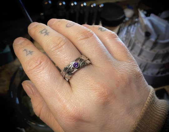 To show scale and size of the completed Amethyst ring, The artist shows what the ring looks like being worn on the middle finger with the tips of the fingers relaxed pointing down. The lighting is slightly dim in the background. The middle of each of the fingers has an astrology symbol tattooed on them. 