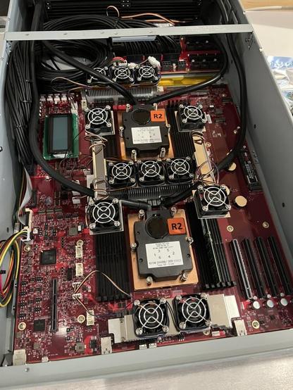 A photo of a motherboard with red silkscreen. There are two large copper heat sinks in the center with flanking banks of 6 DIMM slots on either side of both of the heatsinks. In the very center between the two heatsinks is another set of cooling fins and fans. In the bottom right of the image and motherboard is a bank of PCIe slots. In the top left is a 16x2 character display. There is a reservoir for the liquid cooling at the very top of the image at the back of the chassis.