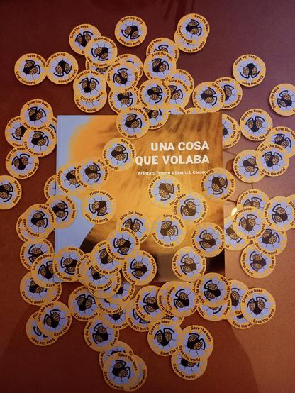 Foto a un ejemplar de "Una cosa que volaba" (escrito por Aránzazu Ferrero e ilustrado por mí) rodeado de pegatinas redondas con el texto "Save the bees, save the world" ("Salva a las abejas, salva al mundo" en inglés) y el dibujo vectorial de la cabeza de una abeja saliendo de una flor azul.