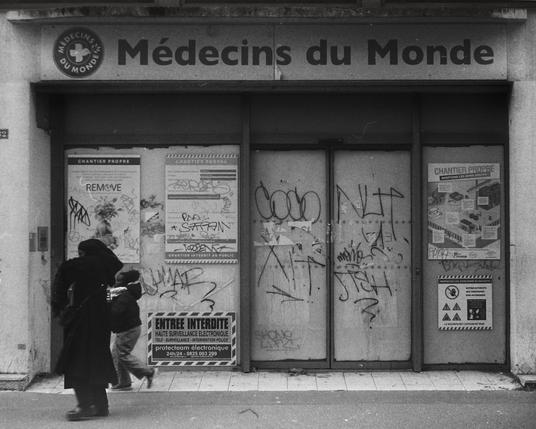 Photo en noir et blanc d'un ancien local Médecins du Monde fermé, ses portes couvertes de graffitis et d'affiches de chantier. Devant, une femme en manteau long marche avec un enfant, capturant un instant de rue chargé d'histoire et de contrastes. Une image qui évoque la précarité, mais aussi le mouvement et l'espoir, parfaite pour rappeler l'importance des actions solidaires comme #Speedon.