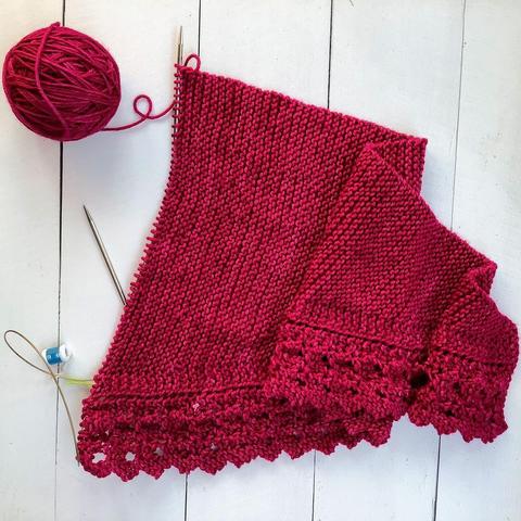 Flat lay view of a ball of dark pink yarn, and a knitting project featuring garter stitch and lace on a circular needle. It's shown on a white plank background.