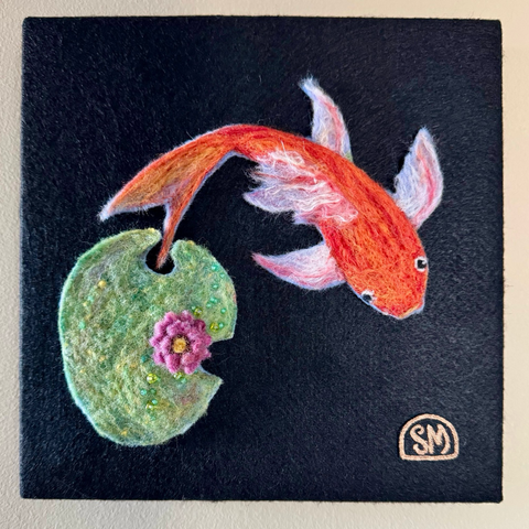 Needle felted orange koi and lily pad with pink flower on black background. Beaded accents on lily pad and metallic embroidery in yellow centre of flower.