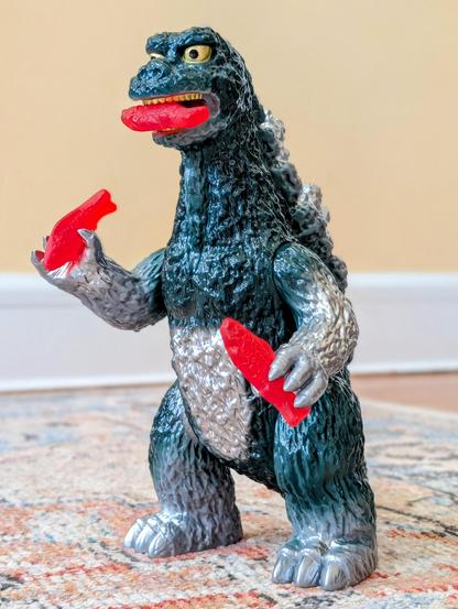 A Japanese vinyl figure of Godzilla. He is holding Swedish Fish candies in both hands, plus there is one in his mouth.