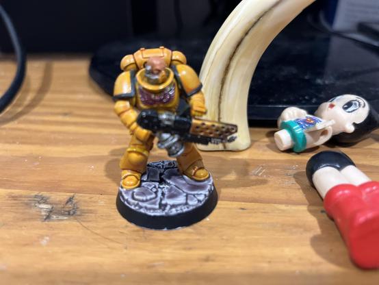 Upon a weathered wooden battleground, a Space Marine warrior stands resolute in gleaming mustard-yellow armour, each panel meticulously highlighted to catch the light like dawn breaking over ancient brass. 

Their battle-worn suit bears the proud heritage of its chapter, with burgundy accents adding depth to the protective plates. In their grip, a heavy bolter speaks of countless battles yet to come. The base beneath their feet tells its tale - a specially crafted terrain of industrial ruins painted in cold metallics that contrast with the warrior's warm armour tones. 

Behind this noble champion, glimpses of everyday life peek into the frame - a curved ivory-coloured form and simple toys that remind us that even mighty warriors have moments of peace. The figurine stands roughly two inches tall, a masterwork of detailed miniature craftsmanship that brings the grim darkness of the far future to vivid life.