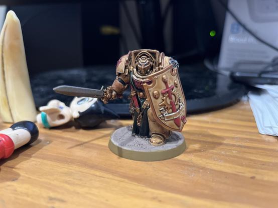 A Terminator-armored warrior emerges from within bronzed ceramite plates, their presence as immovable as a mountain yet as noble as an ancient knight. Their shield, nearly as tall as the warrior themselves, bears proud iconography in deep crimson against weathered gold - a testament to centuries of steadfast service. 


The sword in their grip appears well-worn yet deadly sharp, ready to strike at humanity's foes. The figure stands atop a carefully textured base suggesting urban ruins, painted in subtle greys that ground the warrior's more ornate armour. 


Every detail of the model has been lovingly rendered, from the intricate helm design to the flowing black cloth beneath the armour plates. This figure commands attention at roughly two inches in height through its imposing stance and the obvious care taken in its painting. 