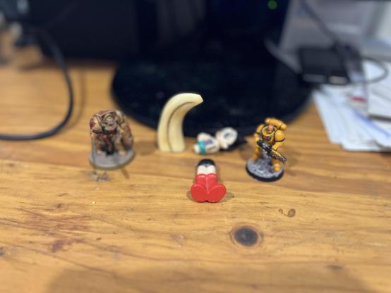 Two champions face each other across a wooden battlefield, their positions suggesting an eternal moment of legendary combat. The mustard-yellow Space Marine stands at the ready with their bolter while their bronze-armoured opponent raises shield and sword in challenge. 


Between them lies a scatter of everyday items - a curved ivory tusk, a memorable token from Africa, its form standing firm on a thin piece of bluetak, now part of the diorama’s landscape where Astro Boy has been ripped apart. One of his cannon arms is missing. My cats and I can only blink and stare, trying to understand the carnage of this mighty confrontation. 


The scene spans roughly six inches across the natural wood grain of what appears to be a desk or table, creating an impromptu arena for these noble warriors. 


The careful positioning and dynamic poses of both figures create a sense of frozen action as if we're witnessing a moment from an epic saga caught between heartbeats.