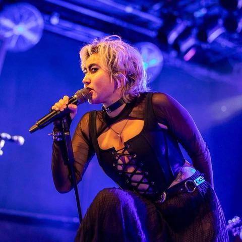Eine Darstellerin mit kurzen, gebleichten Haaren singt in ein Mikrofon, während sie ein schwarzes, geschnürtes Oberteil und einen Choker trägt. Die Umgebung verfügt über eine lebendige Bühnenbeleuchtung mit einem blauen und violetten Hintergrund, der eine energiegeladene Atmosphäre schafft.