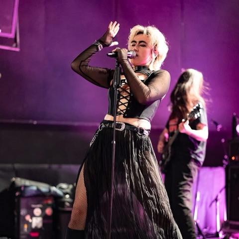 Eine Musikerin tritt leidenschaftlich auf der Bühne auf, trägt ein schwarzes Spitzenoberteil und einen fließenden schwarzen Rock, mit lila Beleuchtung im Hintergrund. Ein Bandmitglied kann hinter ihr gesehen werden, wie sie Gitarre spielt.