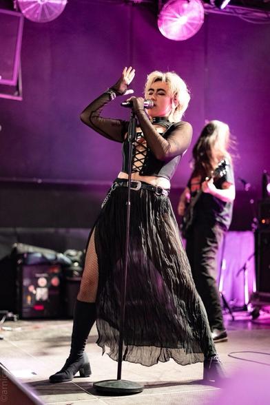A performer sings passionately on stage, wearing a black top with a lace-up design, a flowing sheer skirt, and fishnet stockings. The background features purple lighting, and a guitarist is visible behind the performer.