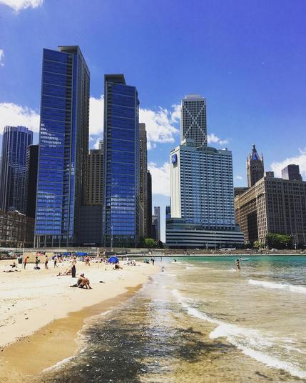 In the foreground is a beachfront with various people sunbathing and playing in the water of Lake Michigan. Beyond them are the skyscrapers of the Streeterville neighborhood of Chicago. 