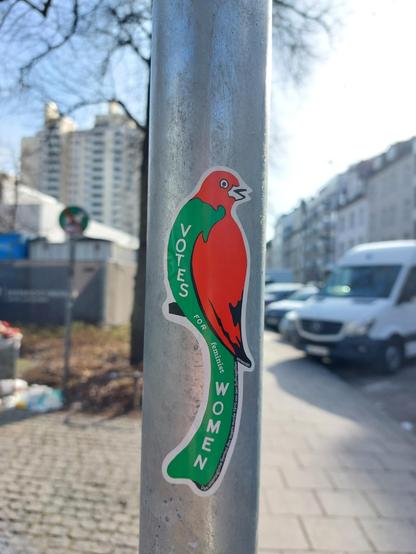 Ein Aufkleber auf der Stange eines Verkehrsschildes mit einem grün-roten Vogel auf dem steht "Votes for feminist Women". 