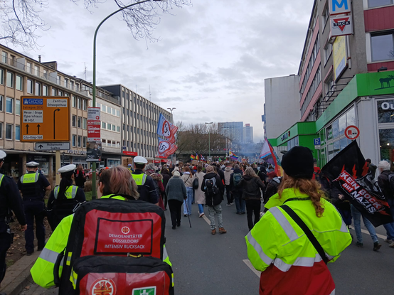 2 Demosanitäter*innen laufen hinter Polizist*innen davor ist die Demo, man sieht Rauch aus einem LKW (Rauchmaschiene) ist zu sehen.