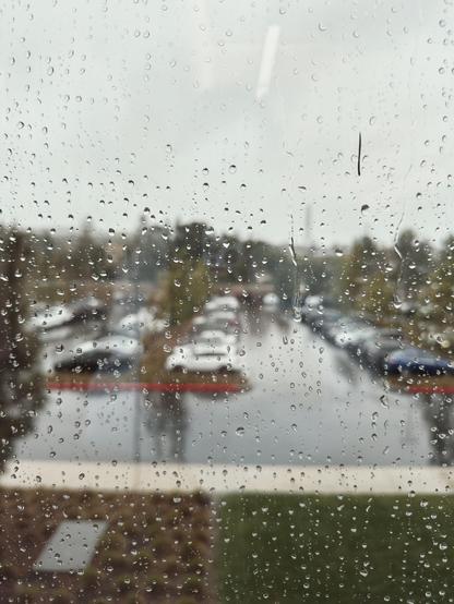 Raindrops on a window with a parking lot and trees blurred in the background.