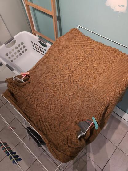 Top view of a dark yellow knitted cardigan lying on a drying rack. The back is sprawled accross, showing the design made with cables of various sizes. Its sides are hanging over the rack's grid . It doesn't have sleeves or a collar yet. 