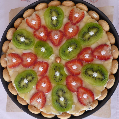 Cake, picture taken from above. It's surrounded by ladyfingers and it has kiwi, strawberry and almond slices on top. In the middle of each fruit slice, there's a snowflake-shaped sugar decoration.