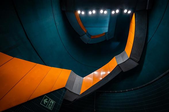 Look-up wide-angle view of a concrete staircase with a bright orange guardrail a peacock blue well at Marienplatz U-Bahn station in München. A trail of little lit lamps on the ceiling is also visible.
