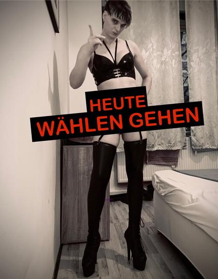Lola, die einen schwarzen Leder-BH, oberschenkelhohe Strümpfe und High Heels trägt, posiert mit strengen Blick und erhobenen Zeigefinger in einem Raum. Die Textüberlagerung lautet "HEUTE WÄHLEN GEHEN".