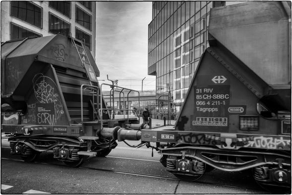 Schwarzweissfotografie zweier Waggons eines Güterzuges; über der Kupplung ist eine Person zu sehen, die vermutlich die Strasse mit den Geleisen überqueren will. 