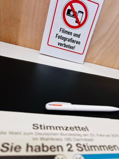 Motiv:
Das Bild zeigt einen Wahltisch mit einem Stimmzettel im Vordergrund, einem weißen Kugelschreiber in der Mitte und einem Hinweisschild im Hintergrund. Das Schild weist mit einem Piktogramm darauf hin, dass Filmen und Fotografieren verboten ist. Der Hintergrund besteht aus einer Holzvertäfelung.

Stimmung & Wirkung:
Die Szene wirkt formell und neutral. Die Unschärfe des Stimmzettels lenkt die Aufmerksamkeit auf das Verbotsschild. Die klare Struktur des Bildes vermittelt Ordnung.

Technische Details:
Bildformat: Dokumentarfotografie.

Farbe: Neutrale Farben mit Schwarz, Weiß und Holztönen.

Schärfentiefe: Geringe Schärfentiefe, Fokus auf dem Schild, unscharfer Vordergrund.

Lichtverhältnisse: Gleichmäßige, künstliche Beleuchtung ohne harte Schatten.