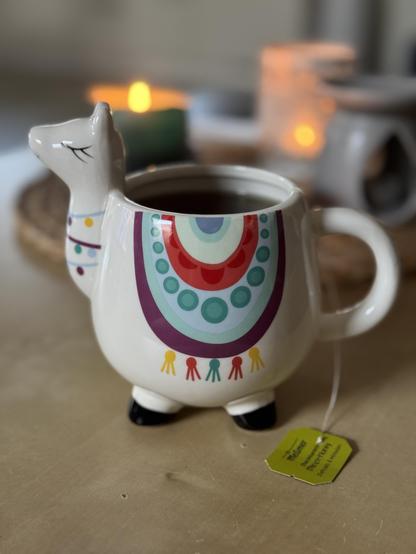 Eine weiße Tasse in Form eines Alpakas mit buntem Schmuck steht auf einem niedrigen Tisch, darin zieht ein Tee. Im Hintergrund unscharf mehrere entzündete Kerzen. 