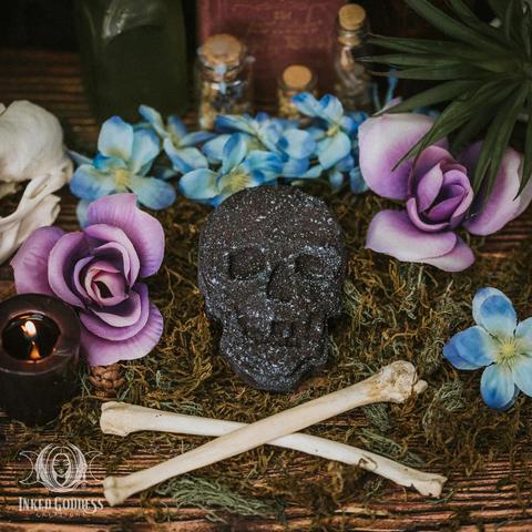 One example of the "Cleansing & Protection Skull Bath Bomb" product is posed on a bed of moss with crossed bones beneath it. The bath bomb is shaped like a flattened skull and is black mottled with lighter material. Presented by Inked Goddess Creations.