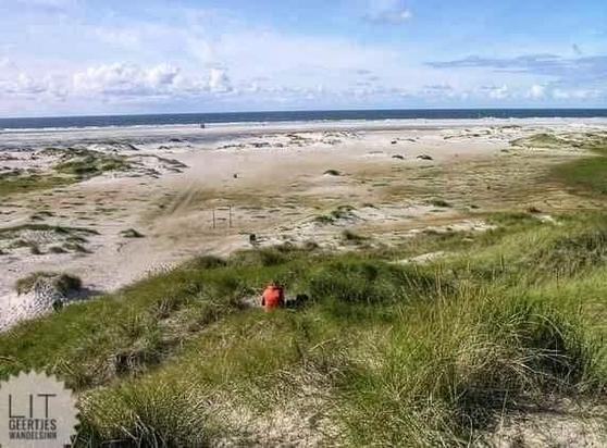 Eine Frau sitzt in den Dünen. Der Strand ist von kleineren Dünen durchzogen. Ein schmaler Wasserstreifen ist zu sehen. Am Horizont kleine Wölkchenbänder.