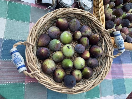 Circular shaped light brown cane basket with blue and white handles contains 24+ figs which are green fleshed & turning purple skinned as they ripen
Edge of photo shows another basket (rectangular shaped) containing more figs
And a row of small glass jars containing brown chutney, they are closed with silver metal clasps and have white label on the lids.
