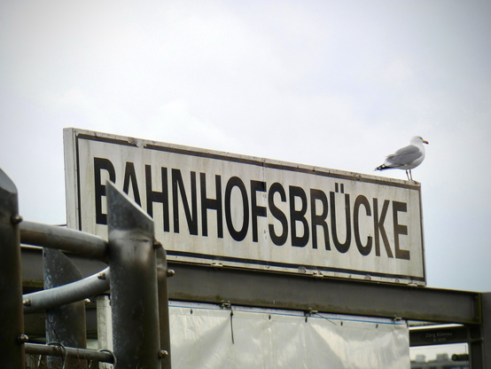 Das Bild zeigt ein Hinweisschild im Kieler Hafen mit der Aufschrift „Bahnhofsbrücke“. Auf dem Schild sitzt eine Möwe. Im Hintergrund ist ein Stück grauer Himmel zu sehen.