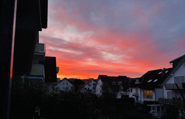 Motiv:
Das Bild zeigt eine Wohnsiedlung mit mehreren Häusern im Vordergrund. Die Dächer und Fassaden sind dunkel, während einige Fenster beleuchtet sind. Der Himmel nimmt den größten Teil des Bildes ein und zeigt eine intensive Färbung mit einer Mischung aus Rot-, Orange- und Blautönen.

Stimmung & Wirkung:
Die Szene wirkt ruhig. Die warmen Farben des Himmels stehen im Kontrast zu den dunklen Häusern. Die vereinzelt beleuchteten Fenster erzeugen einen belebten Eindruck.

Technische Details:
Bildformat: Landschaftsfotografie.

Farbe: Starke Farbkontraste zwischen warmen und kühlen Tönen.

Schärfentiefe: Hohe Schärfentiefe, Häuser und Himmel sind klar erkennbar.

Lichtverhältnisse: Natürliches Licht während des Sonnenuntergangs mit starkem Farbspiel im Himmel.