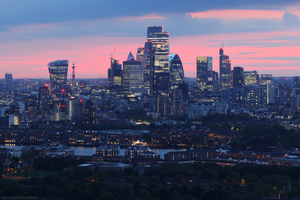 The City of London at dusk. Pink skies with dark blue clouds. Viewed from an elevated location on the Isle of Dogs. River themes in the foreground.