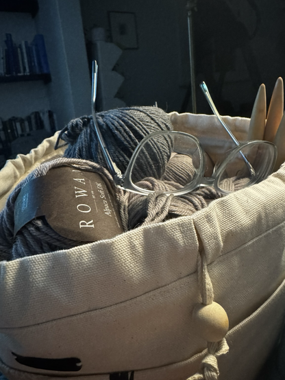 Knitting bag with wool, knitting needles and a pair of glasses resting on the top.