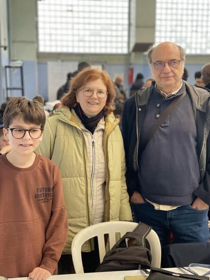 A young boy stands with an older woman and a man in a casual setting. The woman has curly, reddish hair and wears a light green jacket, while the man is in a dark jacket. They are smiling and appear to be enjoying the moment together

Elyes, Maria et l’oncle de Raphaël 