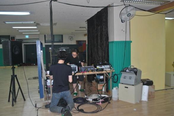 Dans la salle commune du bâtiment municipal, un couple de jeunes gens à cheveux longs vérifie la petite sonorisation, le volume du micro, en tests vocaux « ok, ok, tac, tac, tac, hi, oh, eh, eh, hey, hop, clap, la la la la, aaaaah, ok. ». Ils ont installé les consoles de mixage sur une des tables de ping-pong. Il faut encore brancher les éclairages, mettre du liquide dans la machine à fumée. Un ventilateur suspendu envoie un peu d’air frais, bienvenu, car il fait un peu lourd dans la salle sombre.