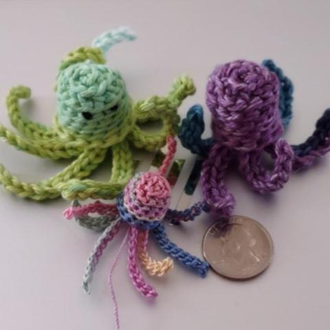 Three small crochetted jellyfish next to a quarter. The two larger jellyfish were made with Peaches and Cream "Stripey" variegated cotton yarn and a 3.125 millimeter crochet hook. The tiny jellyfish was made with pastel variegated crochet thread and a 1.9 millimeter crochet hook.