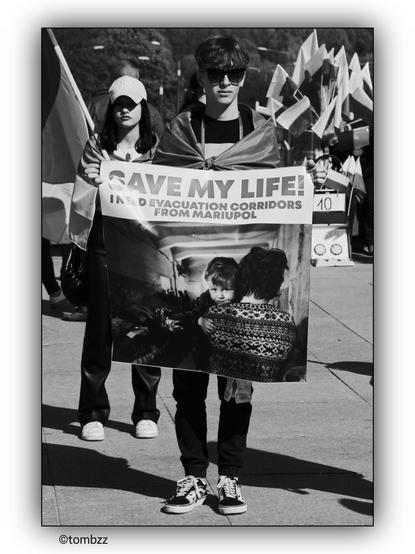 The black and white image shows a young man and a young woman participating in a demonstration. The young man is holding a banner that reads, 'SAVE MY LIFE! I NEED EVACUATION CORRIDORS FROM MARIUPOL.' The banner also features a photograph of a woman holding a child in her arms. The young woman standing next to the young man is holding a Ukrainian flag. In the background, there are more people, some of whom are holding flags. The image is related to the war in Ukraine and the Russian aggression, highlighting the plea for evacuation corridors from Mariupol.