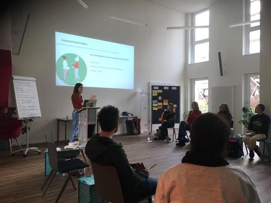 Referentin vor Präsentation mit der Überschrift "Gesprächstechniken". Teilnehmenden davor im Stuhlkreis