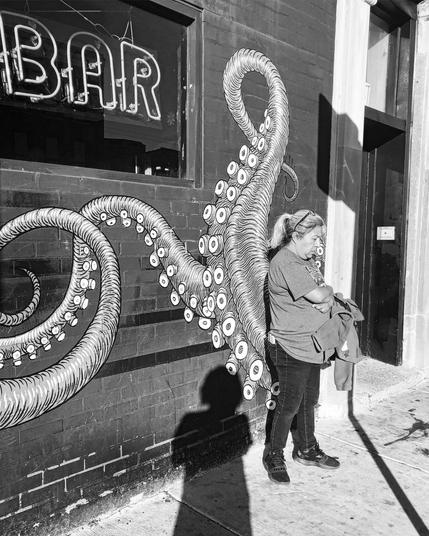 Black and white photo of a woman standing on the sidewalk in front of a mural of an octopus. It appears as if two tentacles are emerging from her back and legs. My shadow bisects the scene (from me taking the picture).
