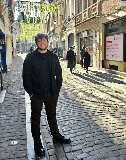 A young man stands in the middle of a narrow cobblestone street in Ghent, Belgium, bathed in sunlight. He’s wearing a black jacket, dark brown pants, and black boots, smiling casually at the camera. The street is lined with older buildings featuring decorative facades; overhead, strands of greenery or hanging decorations cast dappled shadows. A sign on one building reads “Local recherche location sérieuse.” A few pedestrians in dark clothing walk away in the distance.