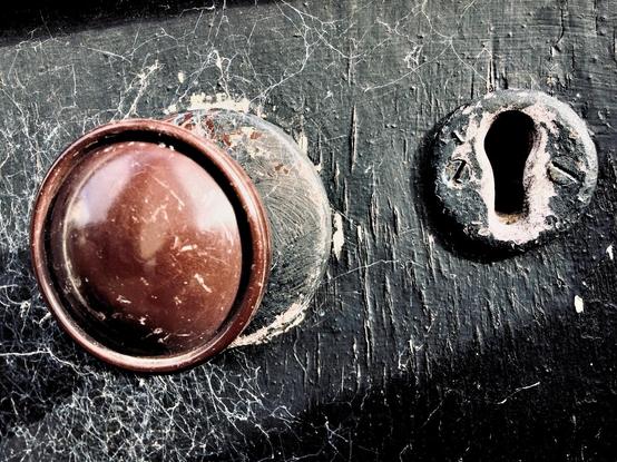 colour photo of brown door knob and keyhole to the right of it. Both are inset in a blank painted wooden door. The paint is pealing whilst revealing the wood grain and spider webs can be seen around the door knob. The knob and keyhole are within a diagonal band of light with shadow above and below.