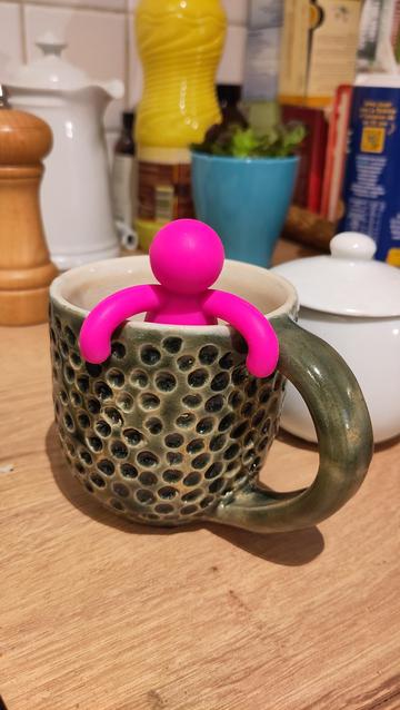 Tassa de ceràmica esmaltada en blanc per dins i en verd ampolla per fora, té textura de foradets. Dins la tassa hi ha un ninot rosa que es repenja a la vora (és un filtre per a infusions).