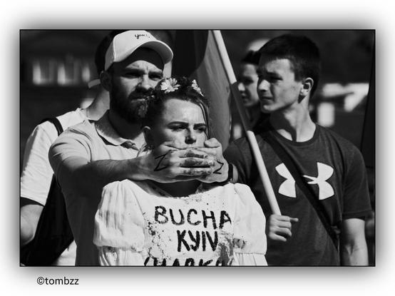The image shows a black and white photograph of a girl with the names of Ukrainian cities: Bucha, Kyiv, and a partially cut-off Kharkiv written on her shirt. A man stands behind her, covering her mouth with his hands. The man's hands have the letter "Z" written on them, a symbol used by Russian invaders to mark their tanks and armored vehicles. His hands are also covered in red paint, symbolizing the crimes committed by Russian soldiers. Next to them, a boy is holding a Ukrainian flag.