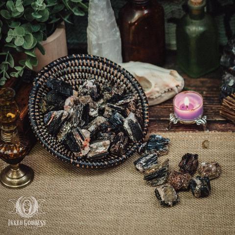 Several specimens of the Black Tourmaline with Mica Raw Piece product are arranged on a burlap-covered surface. Several more specimens fill a tipped grass bowl. They are mostly black columnar tourmaline nuggets with lighter grey and pink mica inclusions on some sides and throughout. Presented by Inked Goddess Creations.
