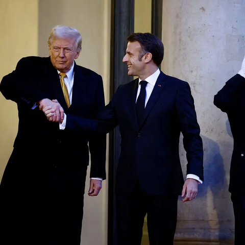 Trump kijkt verbeten en neemt geforceerd, met gekromde arm die hij omhoog moet houden, de bovenhand op een lachende Macron, tijdens een persmoment bij een eerdere ontmoeting.