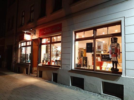Ein Foto der Buchhandlung am Brühl bei Nacht. Man sieht drei hell erleuchtete Schaufenster und das angestrahlte Schild, das rot leuchtet.