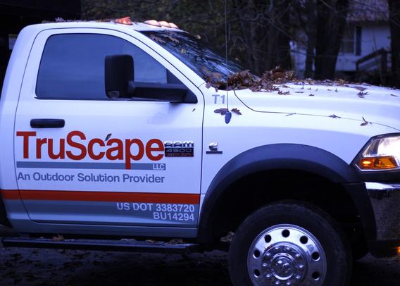 A white TruScape service truck parked with autumn leaves on its hood and roof. The truck door displays "TruScape" in bold red letters with the tagline "An Outdoor Solution Provider" underneath. US DOT and registration numbers are visible on the lower door panel. The truck appears to be a heavy-duty commercial vehicle with industrial wheels, photographed in the fall with trees in the background.​​​​​​​​​​​​​​​​