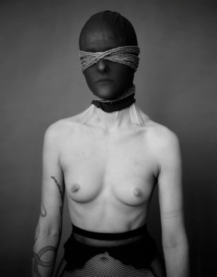 Foto a mezzafigura a modella in topless con una calza in testa e una corda che stringe al collo e sugli occhi.