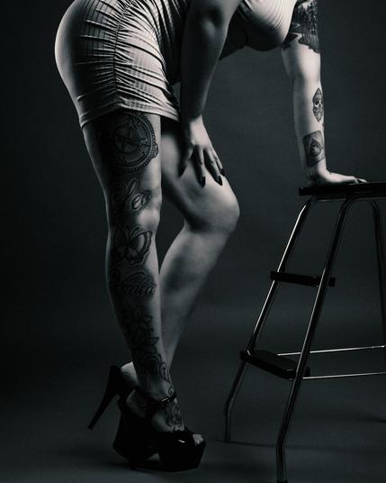 A black and white studio portrait of a tattooed woman wearing a tight dress and high heels, leaning against a metal stool. The composition highlights her leg tattoos, including a compass, butterflies, and script, with a dramatic lighting setup emphasizing the contrast.