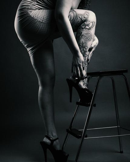 
ALT Text:
A black and white studio portrait of a tattooed woman wearing a tight dress and high heels, leaning against a metal stool. The composition highlights her leg tattoos, including a compass, butterflies, and script, with a dramatic lighting setup emphasizing the contrast.